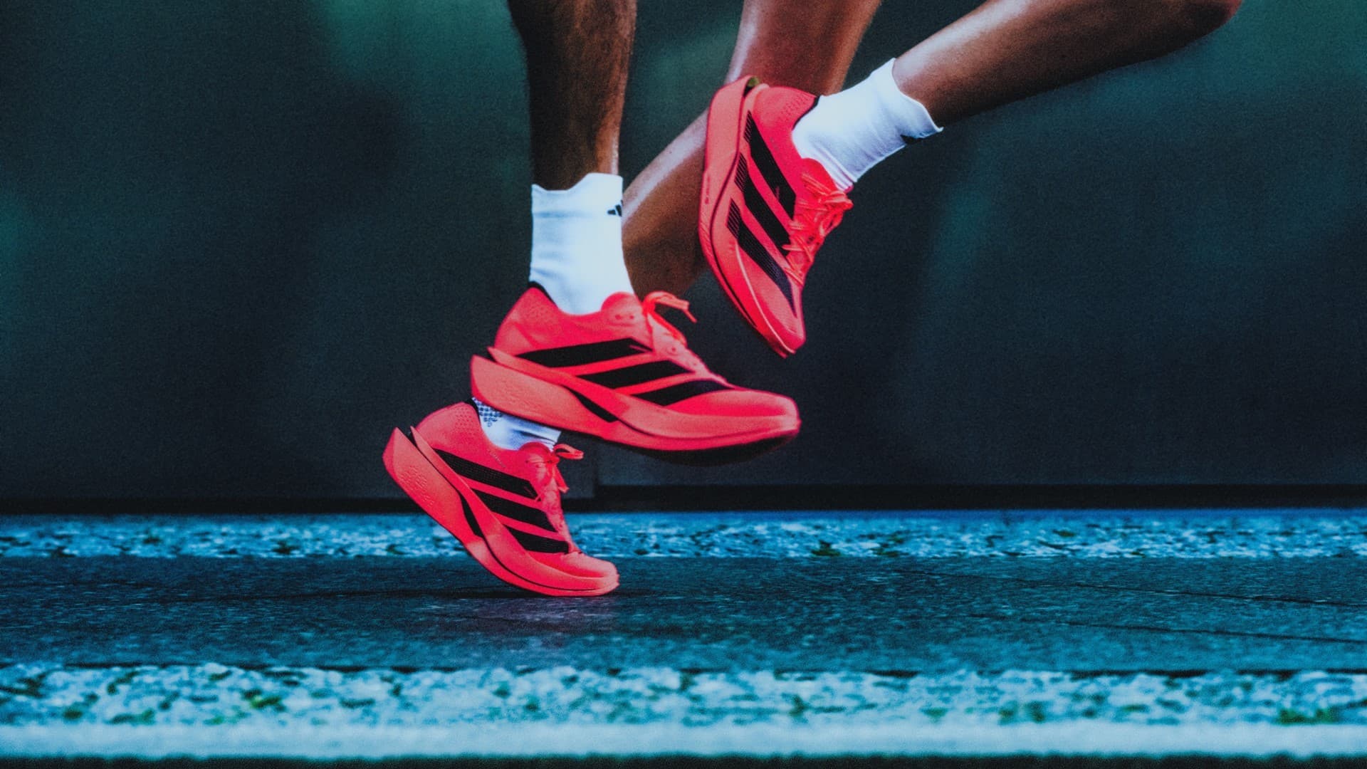 Pés de corredor em suspensão sobre o asfalto, calçando o adidas Adizero Adios Pro 5 vermelho com três listras pretas