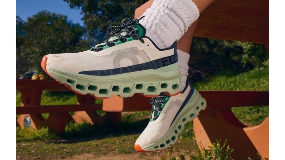 Par de tênis On Cloudmonster branco e verde com solado CloudTec, calçado por uma corredora sentada em um banco