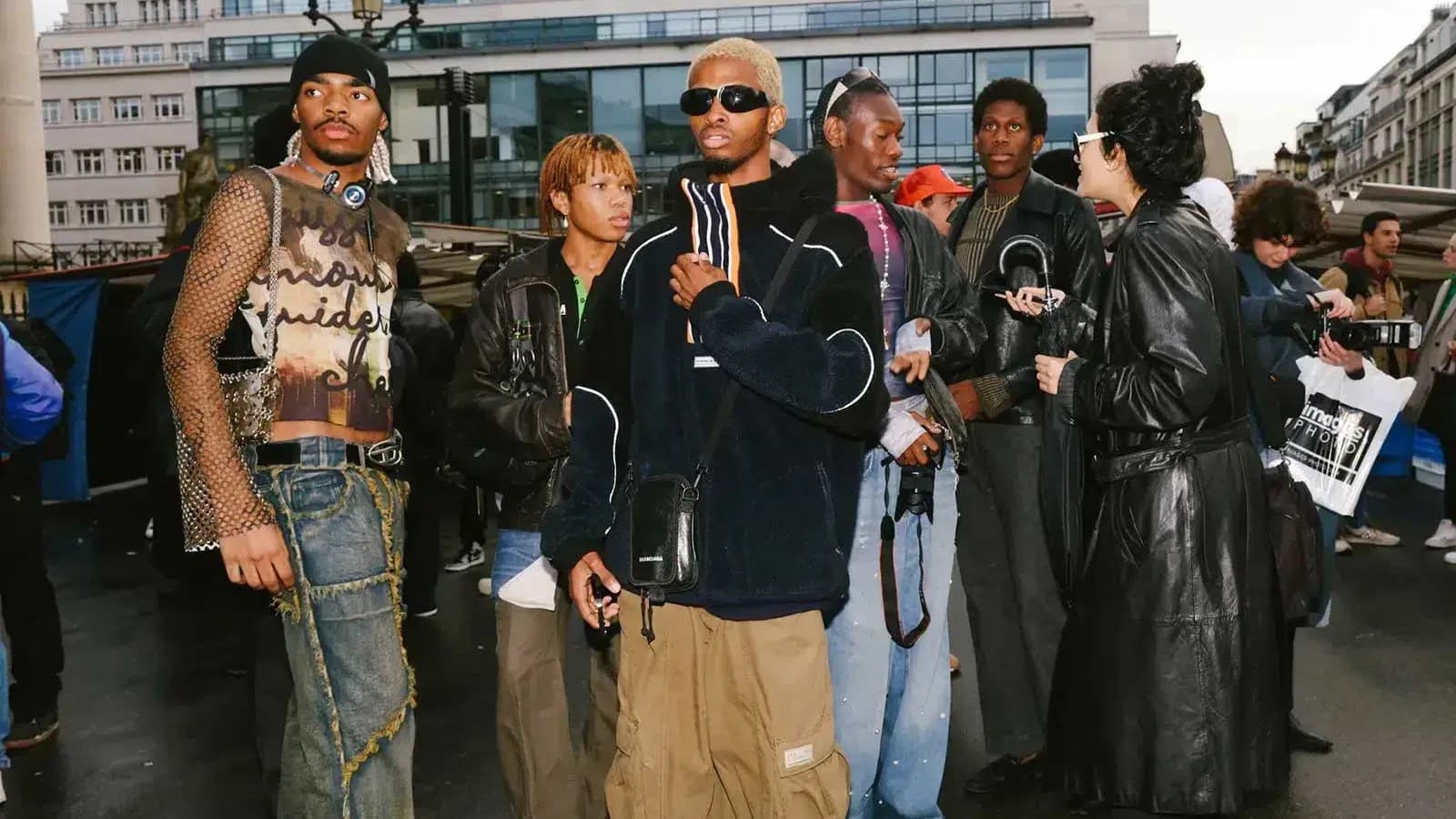 Grupo de jovens em roupas de streetwear reunido em uma rua de calçamento molhado, com jeans largos, jaquetas de couro e bolsas a tiracolo