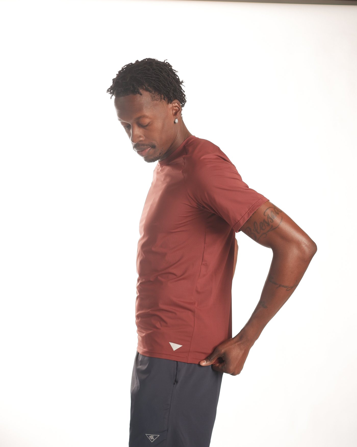 Homem de camiseta vinho e bermuda azul-marinho da Studio Athletic Club, em pé sobre fundo branco de estúdio